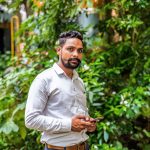 A handsome young man standing outdoors among lush greenery, holding a smartphone.
